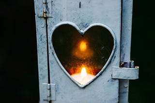 Gray metal hinged door with heart shape cut out of the middle with a lit candle showing.