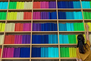 A person is reaching up high for a book on a bookshelf. There are multiple bookshelves beside this one, with hundreds of multi-coloured books on all of them.