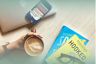 A person holding a coffee while at their desk. On the desk is a smart phone and two business books.