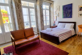 The inside of a studio apartment in Braga with granite walls, three French doors with windows, a small loveseat, a bed and a table and chair.