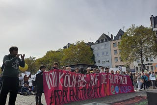 A magenta FFF Cologne banner “From Cologne to the Philippines” held up by local schoolchildren under grey skies on the Cologne Heumarkt for the Global Climate Strike 23.9.2022. The banner names the cooperation with YACAP — “Youth Advocates for Climate Action Philippines” and the hashtag #PeopleNotProfit, in the foreground and background grown-up attendants of the climate strike show their support with applause and signs.