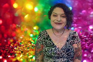 A glittery rainbow color background contains an image of Bela on the right. She is a light-skinned Latina with short, dark, curly hair and blue green eyes. She is wearing a black and white dress and has tattooed sleeves.