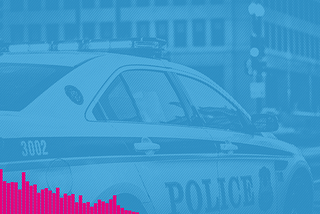 A view of the passenger side of a police car as it drives down a city street. A large building is in the background.