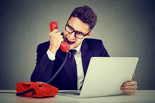 Man at desk in front of laptop yelling into a red land line.