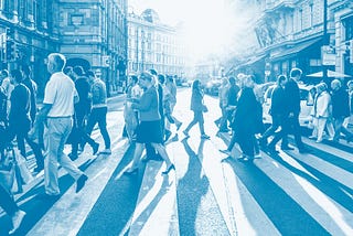 Photograph shows a lot of different types of people walking across a main road crossing using a light blue duotone