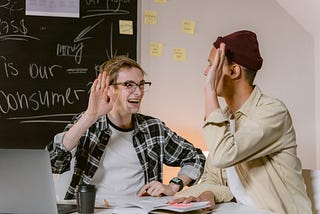 Two men sitting on a table with open books and about to high-five.