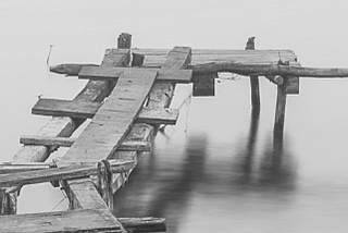 A black and white photo of a ramshackle dock that extends into a featureless lake