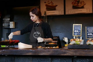 Photo Description: woman in gray apron preps food behind a bar.