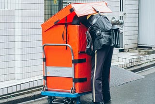 postman in a gray uniform is looking inside a large orange delivery cart, which is partially open, near a white tiled building on a city sidewalk.