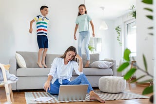 Tired young mother sitting on floor with laptop while naughty children jump on sofa