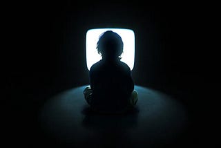 A kid sitting in front of a tv screen with flashing light in a dark room. He is so focused on it as he lose track of time he spents on it.