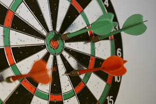 A dartboard with a lot of holes and four darts fixed at it with one right at the target. The dartboard represents a machine learning model and the darts are hyperparameters. The one at the target is a hyperparameter with best value that gives the highest accuracy of the model.