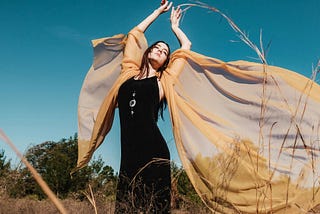 A young person with long dark hair stands with their arms held high in a field with a translucent shawl billowing around in the light breeze like wings