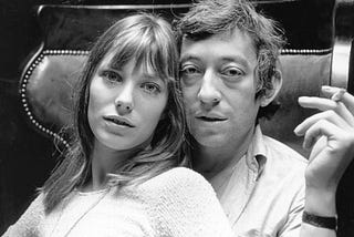 A picture of Serge Gainsbourg with his romantic partner sitting on his lap. Serge Gainsbourg holds a cigarette in his left hand.