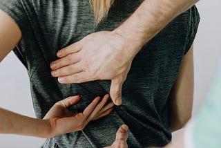 A person is seated with their back in view and motioning toward their left side with their left hand while in the right a second person is reaching both of their hands towards the back as if they are going to massage or adjust that area of the person’s back.