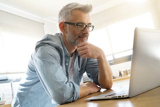 A grey-haired man wearing glasses smiles slightly at his computer screen.