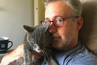 A man wearing red glasses snuggles with a gray cat.