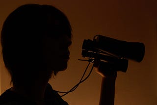 A person with shoulder length hair lit from behind, appearing like a shadow, holding a pair of binoculars
