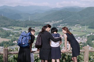 4 young women who are sisters in an embrace looking upon a vast spread of mountains and grasslands.