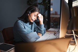 Person sitting at a desk with a headset, eyes closed, holding a tissue to their forehead, looking exhausted while working at a computer.