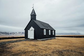 An antique black church with white doors and windows on open land surrounded by mountains in Iceland.