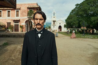 Colonel Aureliano Buendía stands in the town square of Macondo, wearing formal 19th-century black attire, with colonial buildings and villagers in the background. Scene from Netflix’s One Hundred Years of Solitude series.