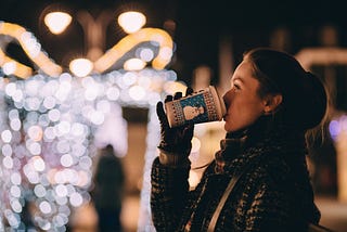 Pleasant brown haired tanned caucasian woman drinking takeout coffee on street corner at night with festive clear white winter lights