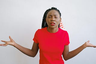 A black woman in a red t-shirt closes her eyes in exasperation and shrugs, arms outstretched.