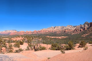 Distant mesas contrast against the bright blue sky and in the foreground a few tiny figures stand on large, round surfaces of red rock