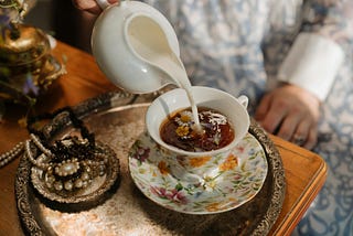 Image by cottonbro studio — Pexels. A close-up image of a woman’s hand pouring milk from a white teapot into a delicate floral teacup filled with tea and chamomile flowers. The scene is set on a silver tray ornamented with elegant pearls, creating a soft and cozy vibe.