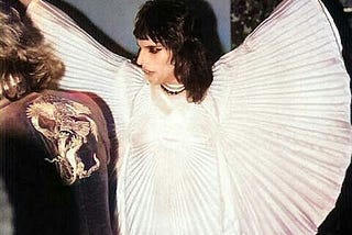 Freddie Mercury wearing a ruffled, white cloak in a slightly dim room.
