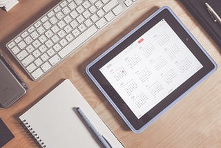 A desktop with a computer keyboard, notepad and pen, and iPad with the calendar pulled up on the screen.