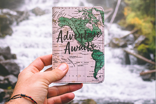 A man’s hand holding a card that says ‘Adventure Awaits’ against the backdrop of a waterfall.