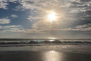 Image of a beach with ocean waves crashing. The sky is blue and the sun is shining through the clouds.