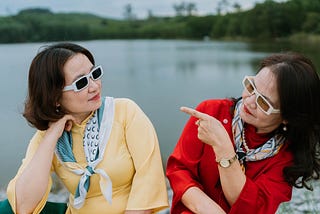 Hey middle age? You look fabulous. Two women in silly sunglasses and cute sweaters glam it up by a lake.