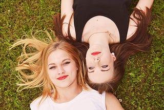 A blonde and a brunette with red lipstick lying down on grass.