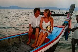 A tanned white couple sit in longtail boat painted blue with red edging. She is wearing a white dress and holding a bouquet and he is wearing a white shirt and pinstriped shorts. A Thai fisherman is holding onto the bow.