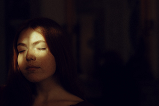 An image of a young lady standing in dark room with a thin sunlight beam shining from the left side of her face
