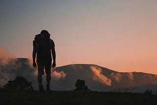 A silhouette of a person trekking towards an orange sky and large mountains.