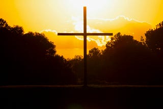 A brown cross above the water in front of a yellow sky.