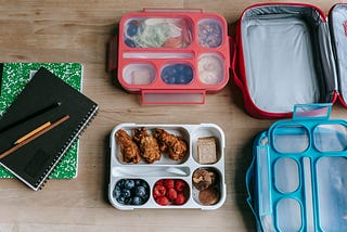 Photo of pink and blue lunch boxes and trays on table next to a green and black notebook and pencils.
