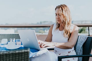 Image of a woman working on her laptop at a table on a balcony overlooking a cityscape