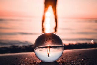 A figure standing on a glass ball as the sun sets on the beach