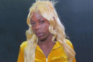 head and shoulders photo of a black woman wearing a gold blouse. she has long blond hair past her shoulders. She is not smiling, but she is looking at the camera