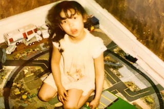Little girl with dark hair sitting in the middle of a train set looking up at the camera.