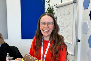 A picture of Ruth Baughan, Lead Service Designer at Boots, wearing glasses and a red sweater smiles while holding a caterpillar-shaped chocolate cake in an office meeting room.