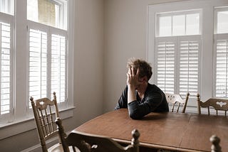 A man is sitting at a kitchen table. He is holding his face in his hand with his head turned toward a window.