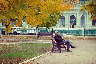 An elderly man sitting on a park bench. He is wearing an overcoat and holding a walking stick. The yellow and gold leaves of two trees are visible in shot, and fallen leaves litter the ground. There are no other people visible in the park.
