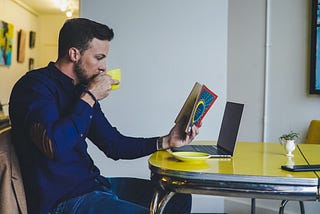 A man reading a book at a yellow table while drinking from a yellow cup.