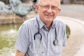 A man with glasses and blue striped shirt with stethoscope around his neck sits on the side of a water fountain, smiling at the camera